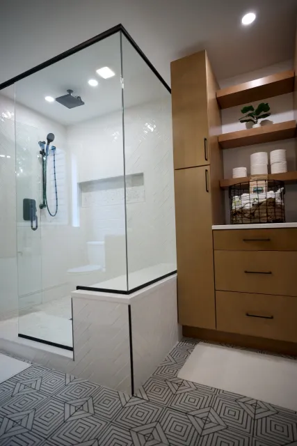 Modern bathroom with a glass shower enclosure, white herringbone tile walls, wooden shelving with towels and a plant, patterned tile floor, beige cabinetry, and ceiling lights.