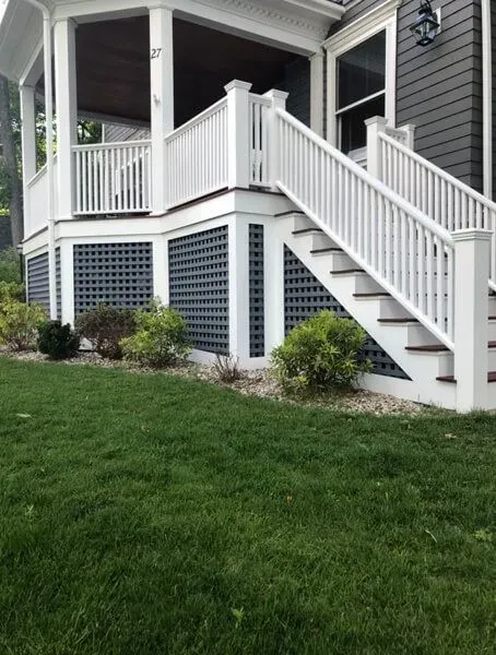 White porch with stairs, lattice skirting, and small bushes on green lawn.