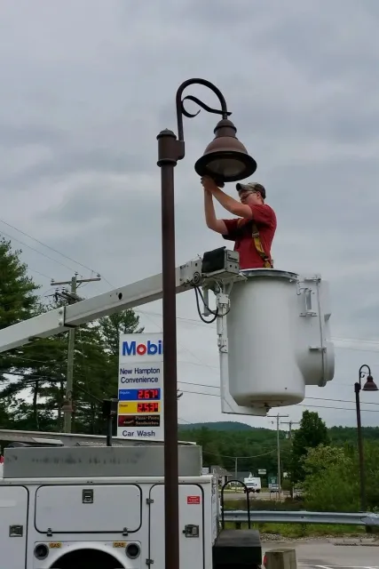 A worker in a cherry picker adjusting a street lamp in a parking lot near a gas station with a sign displaying fuel prices.