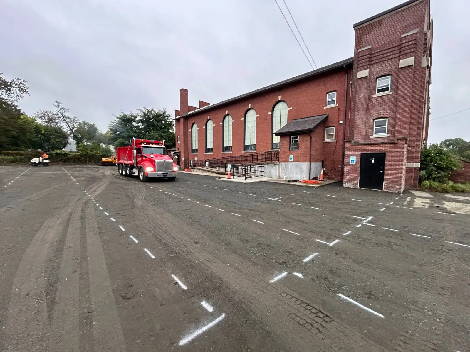 A red dump truck idling in a gravel parking lot next to a brick building with white markings on the ground for layout.