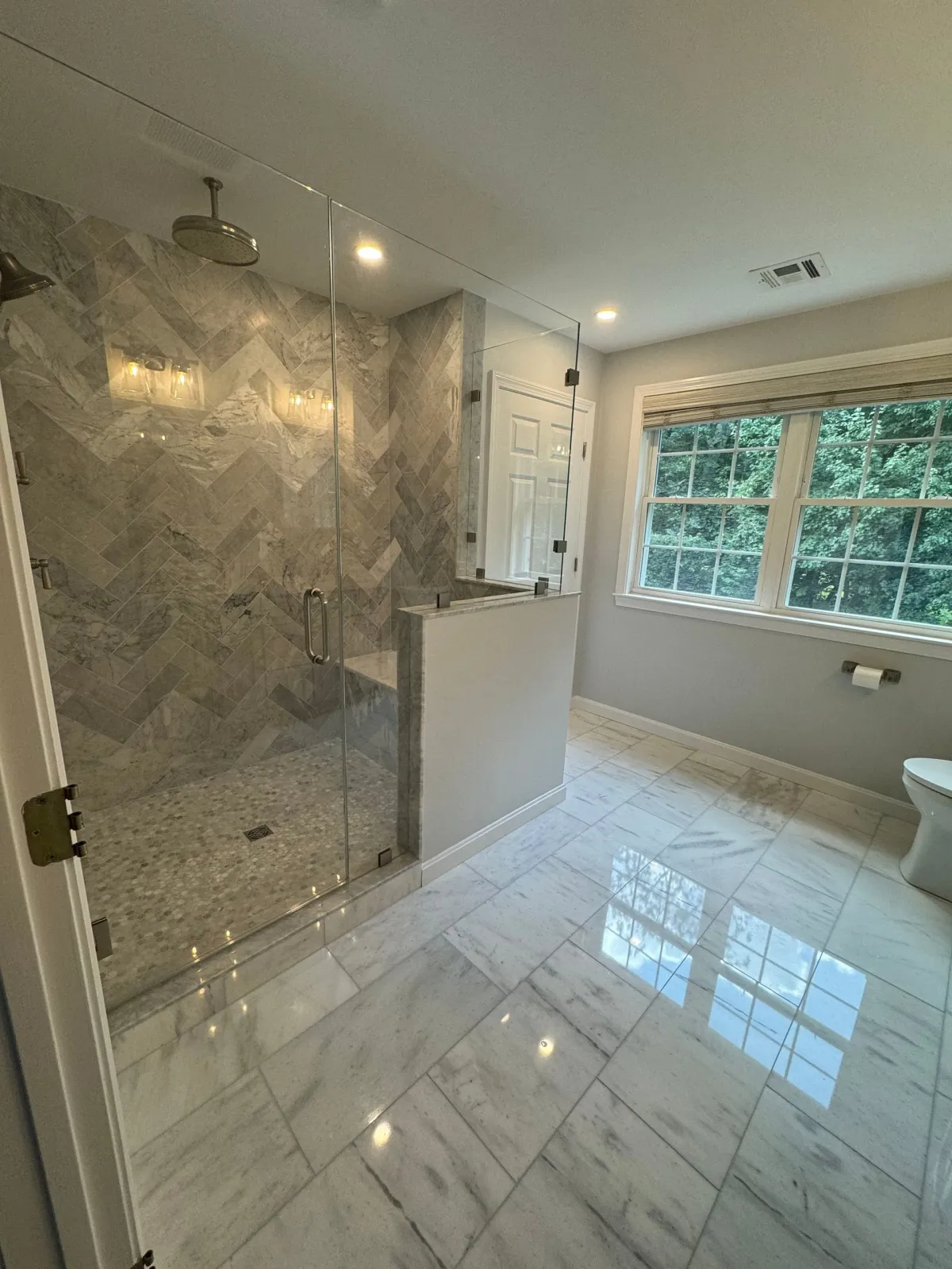 A modern bathroom with marble-tiled floors, a walk-in shower with chevron-patterned walls, and a large window.