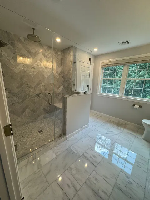 A modern bathroom with marble-tiled floors, a walk-in shower with chevron-patterned walls, and a large window.
