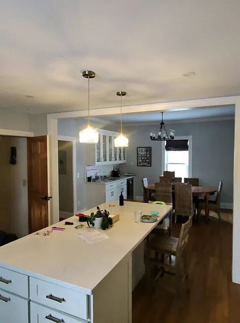Wide view of a residential kitchen and dining area with a white kitchen island, pendant lights, and hardwood floors in Wellesley, Massachusetts.