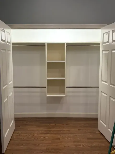 A closet with white doors, hardwood floors, two clothing rods, and a center shelving unit with three open compartments.