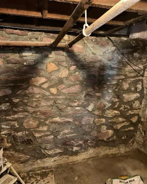 A basement wall constructed of irregular fieldstone and mortar, viewed from a low angle with overhead pipes.