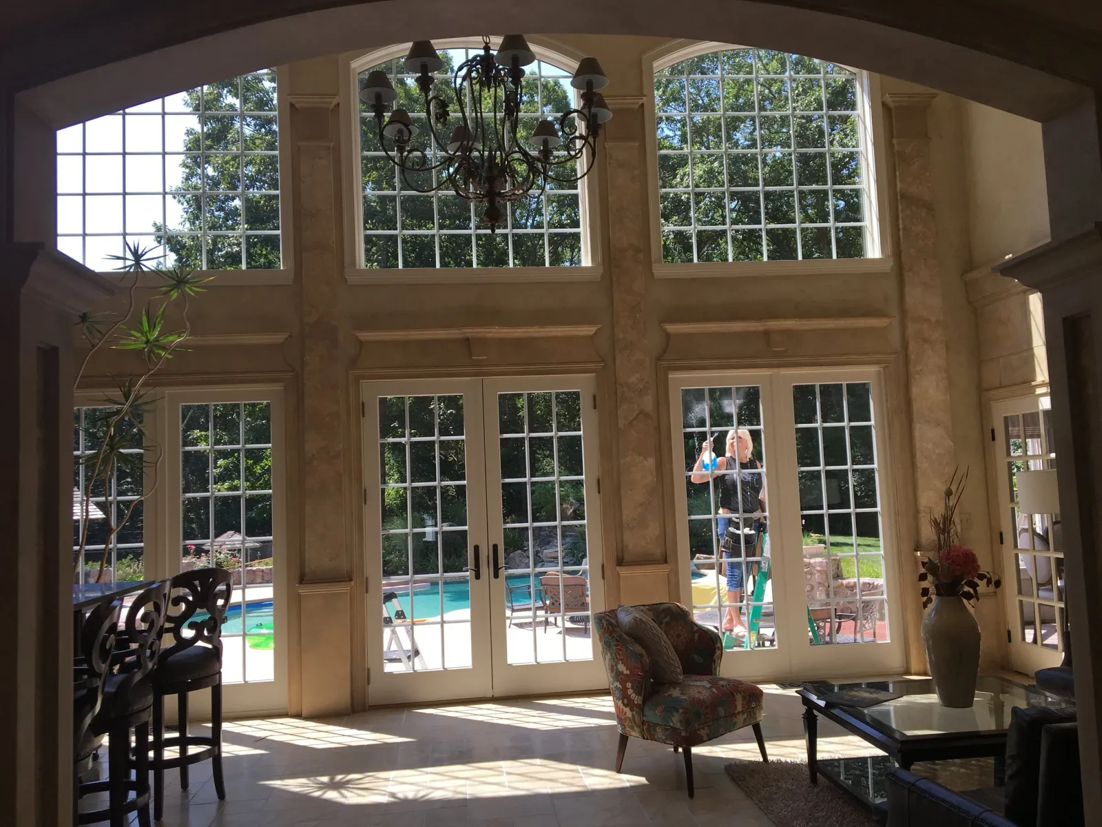 Interior room with large windows and garden view, featuring a chandelier, chairs, and a table. Outside, a person is cleaning the windows near a swimming pool and trees.