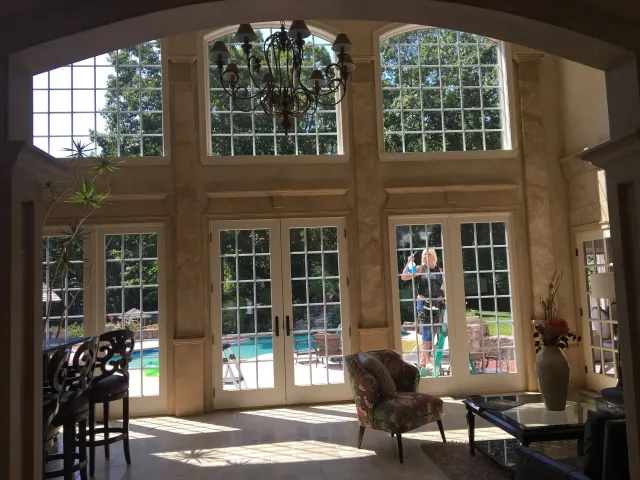 Interior room with large windows and garden view, featuring a chandelier, chairs, and a table. Outside, a person is cleaning the windows near a swimming pool and trees.