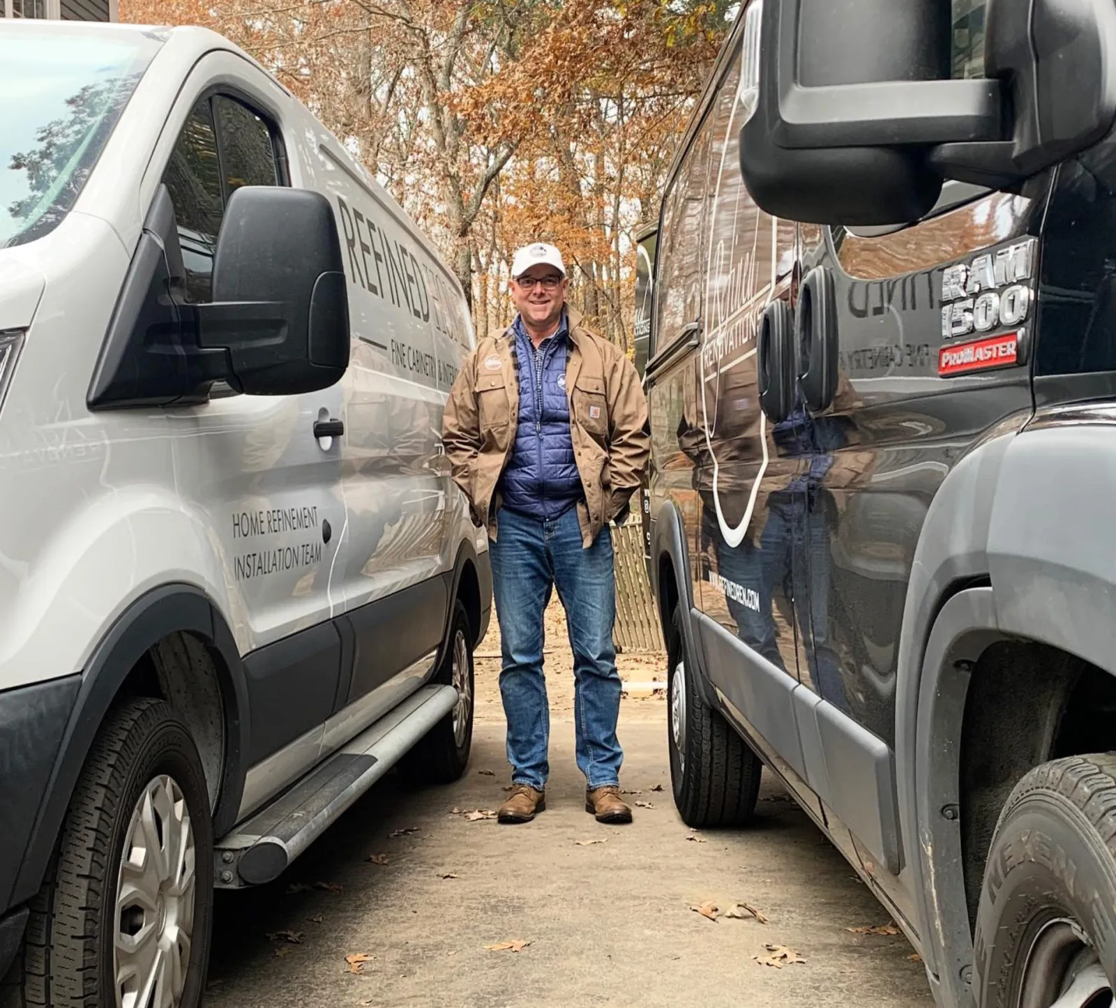 Jim Buhrer, owner of Refined Renovations, stands between two vans wearing a cap and a beige jacket with jeans.