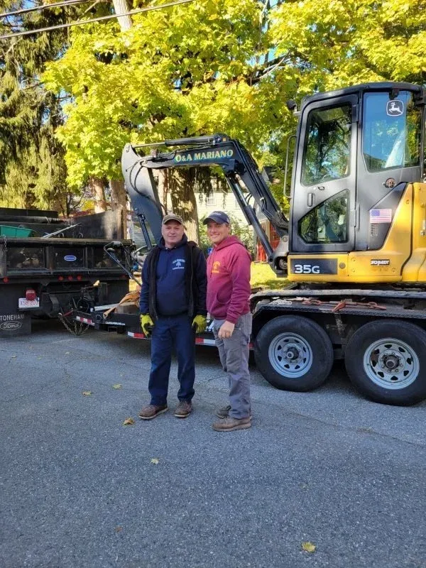 Two men standing in front of a yellow John Deere excavator on a flatbed truck, with trees and a blue sky in the background.