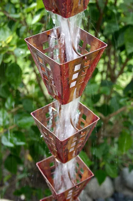 A rain chain with four stacked square containers, water flowing down through each, outdoors with green foliage in the background.
