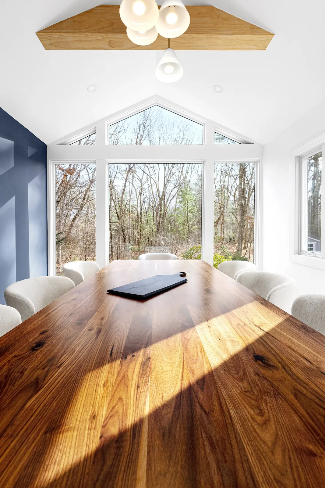 A bright dining room with a large wooden table and white chairs, big windows showing a wooded outdoor view, a ceiling light fixture, and a dark blue accent wall.