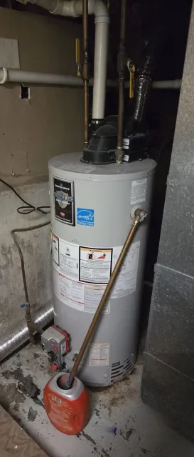 A water heater installed in a basement with pipes, vents, and an extension cord nearby, along with a red container. The water heater has energy efficiency labels and safety warnings.