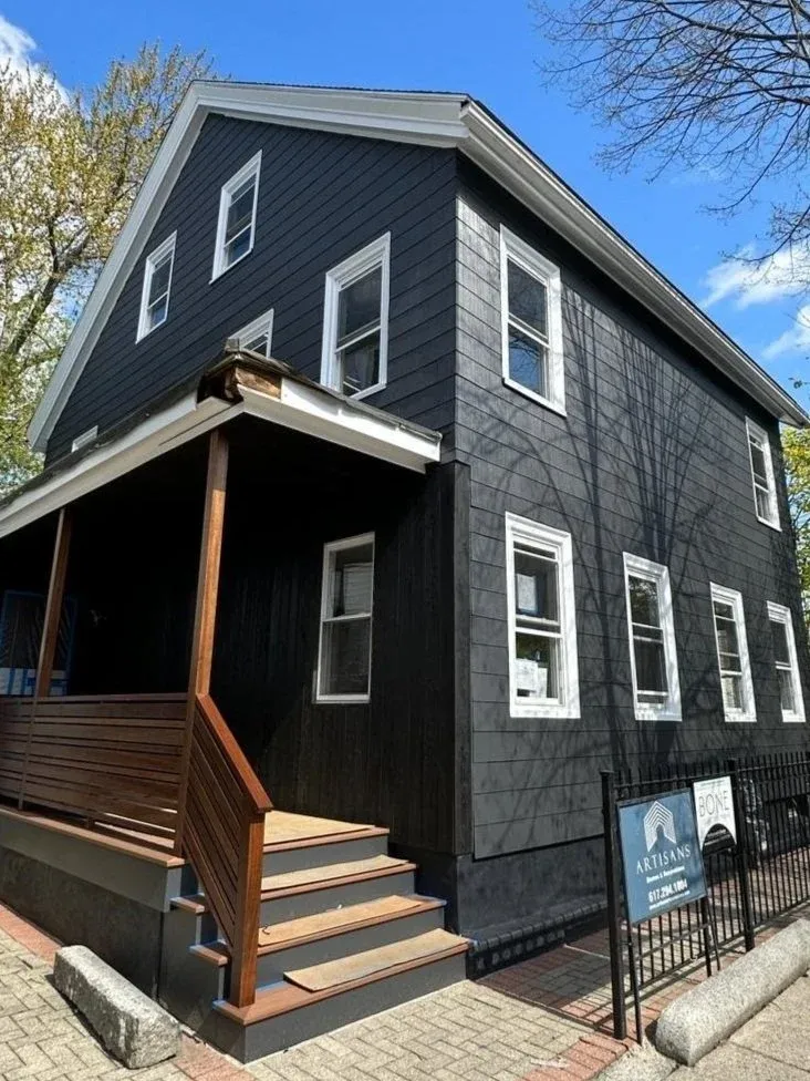 A two-story black house with white window frames, front stairs with wooden railings, and a small covered porch area, under a sunny blue sky.