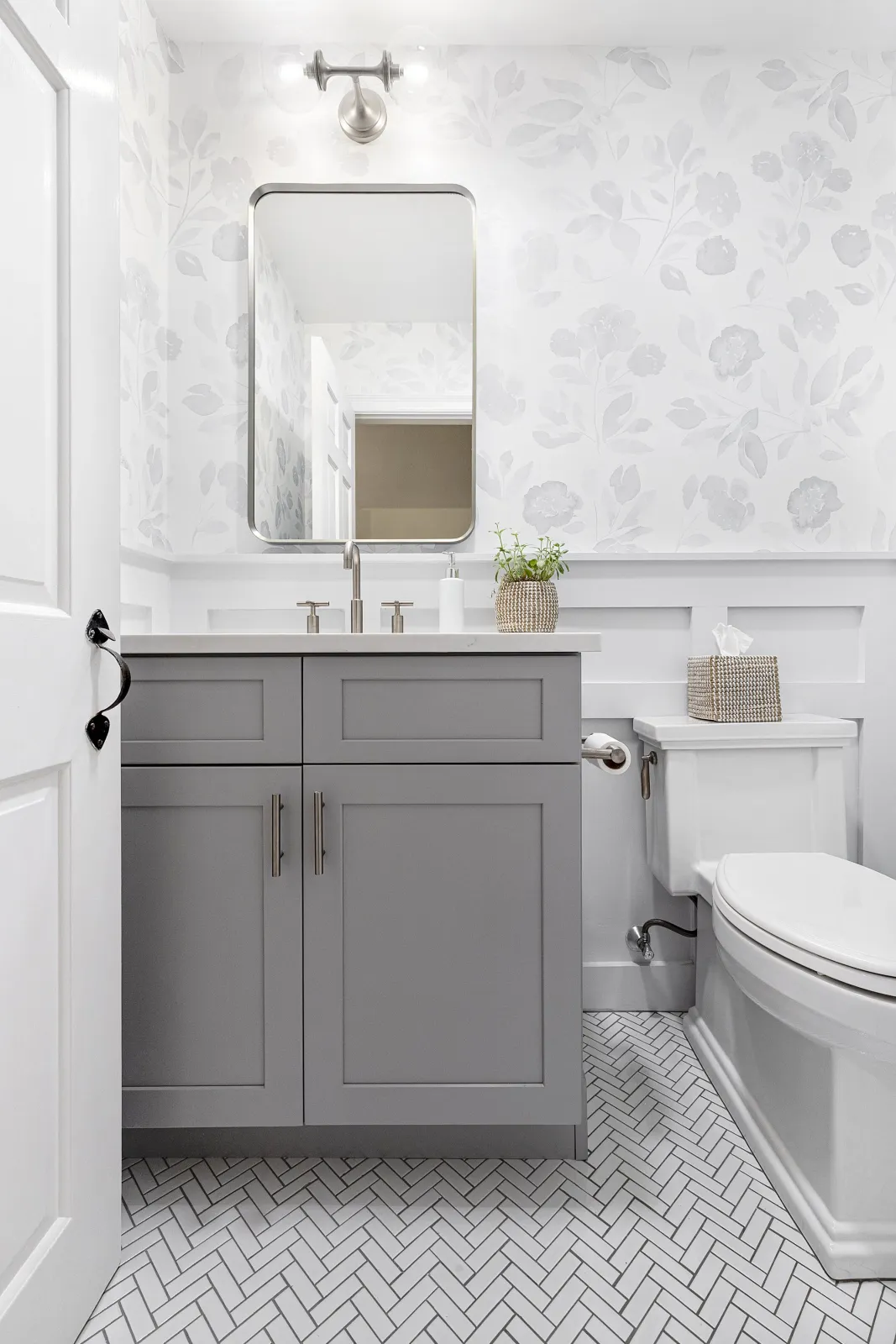 Bathroom with gray vanity, white walls with floral wallpaper, rectangular mirror, white toilet with tissue box, small potted plant, and patterned tile floor.