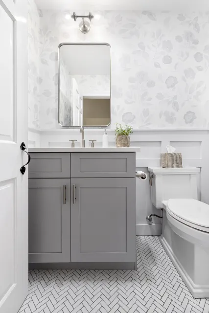 Bathroom with gray vanity, white walls with floral wallpaper, rectangular mirror, white toilet with tissue box, small potted plant, and patterned tile floor.