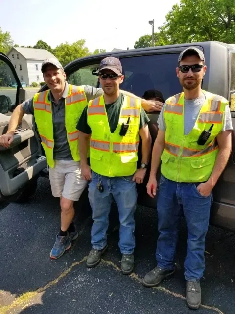 Three men wearing reflective safety vests standing next to a large vehicle on a paved surface with houses and trees in the background.