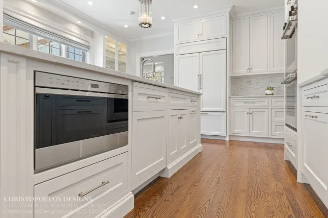 A modern kitchen featuring white cabinetry, stainless steel appliances, a marble countertop, hardwood flooring, and a small potted plant on the counter. Large windows with blinds let in natural light.