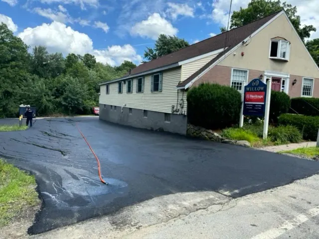 Asphalt parking lot being sealed next to a building on a sunny day. A worker is seen applying the seal.