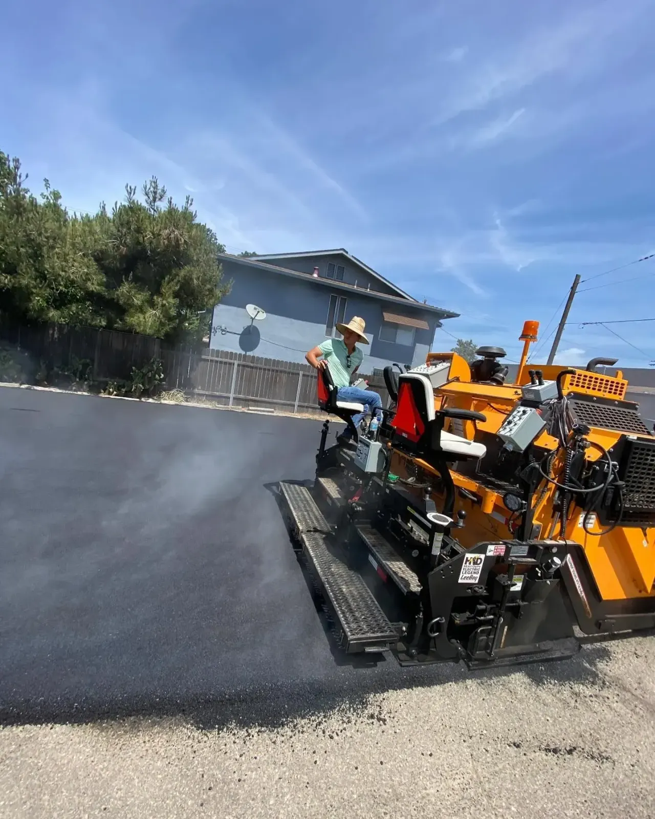 Person operating asphalt paving machine on a road under a blue sky.