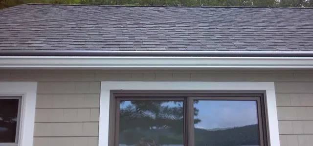 A tan house exterior with white window trim, a horizontal window, and a grey shingled roof under a gutter.