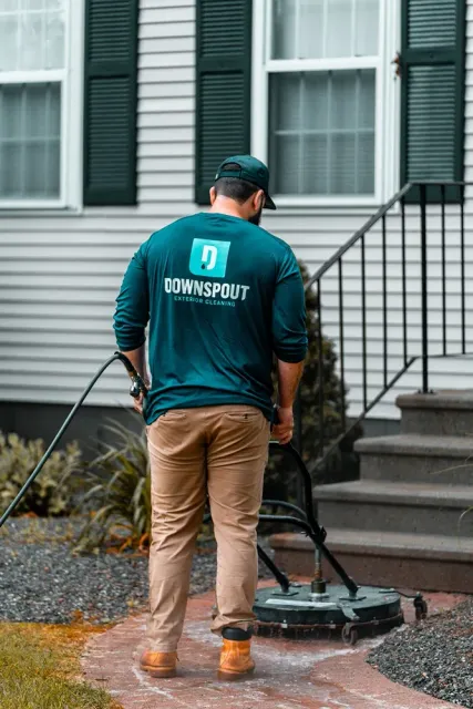 Worker wearing a Downspout Exterior Cleaning shirt using a surface cleaner on a brick walkway in front of a house.