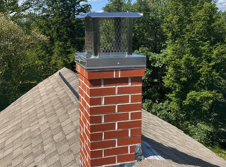Close-up of a brick chimney with a metal cap and a mesh guard on a residential roof, surrounded by green trees.