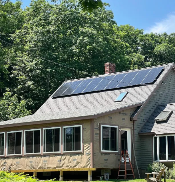 A house under renovation with new solar panels installed on the roof, surrounded by green trees under a clear blue sky.