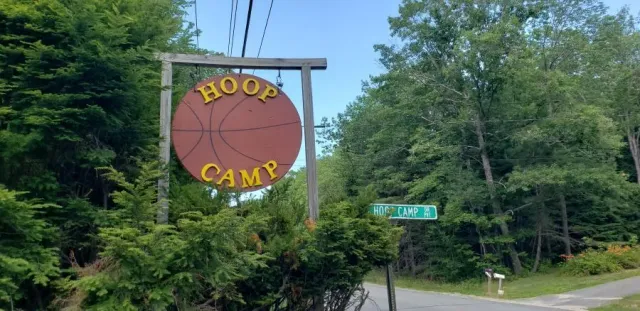 A sign for a hoop camp is hanging from a pole in the middle of a forest.