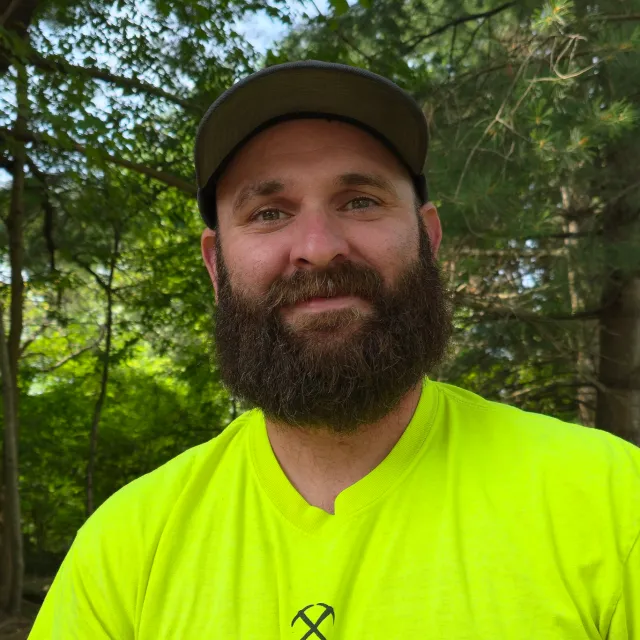 A man with a beard wearing a neon yellow shirt and a cap outdoors among green trees.