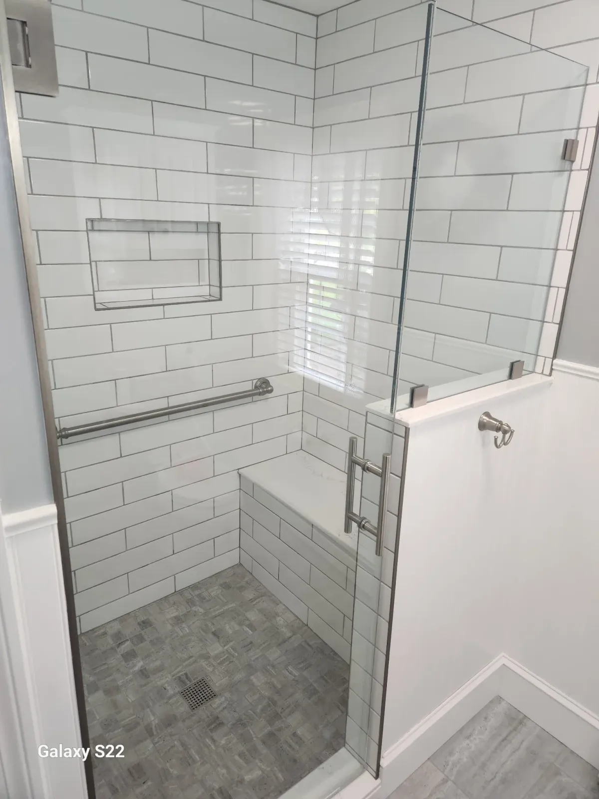 A modern shower with white subway tiles, a built-in bench, a grab bar, a recessed shelf, and a glass enclosure with metal hinges and handle.