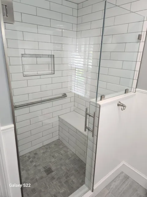 A modern shower with white subway tiles, a built-in bench, a grab bar, a recessed shelf, and a glass enclosure with metal hinges and handle.
