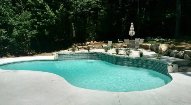 Backyard swimming pool with patio furniture and an umbrella, surrounded by trees and rocks.