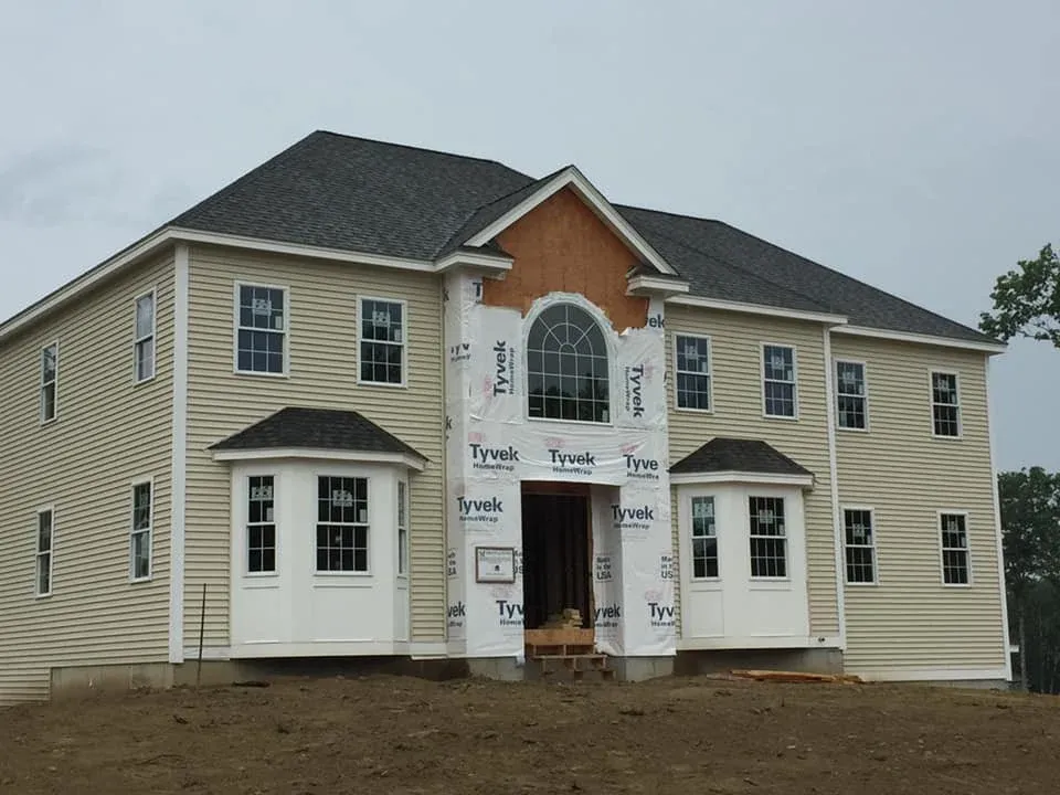 Two-story house under construction with siding partially installed and a large window above the entrance, with fresh soil in the front yard, cloudy sky in the background.