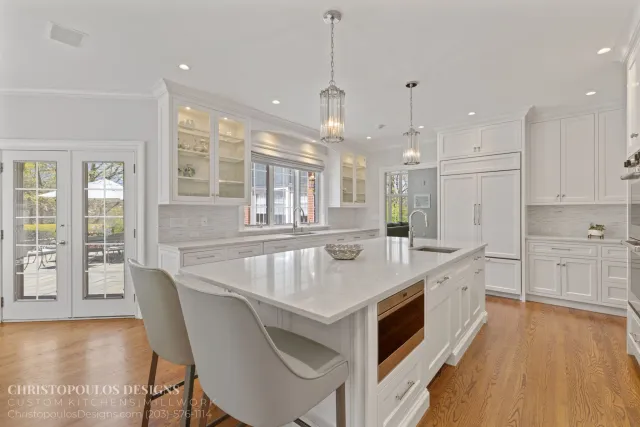 Bright, white kitchen with an island, pendant lighting, and large windows and glass doors showing an outdoor patio area.