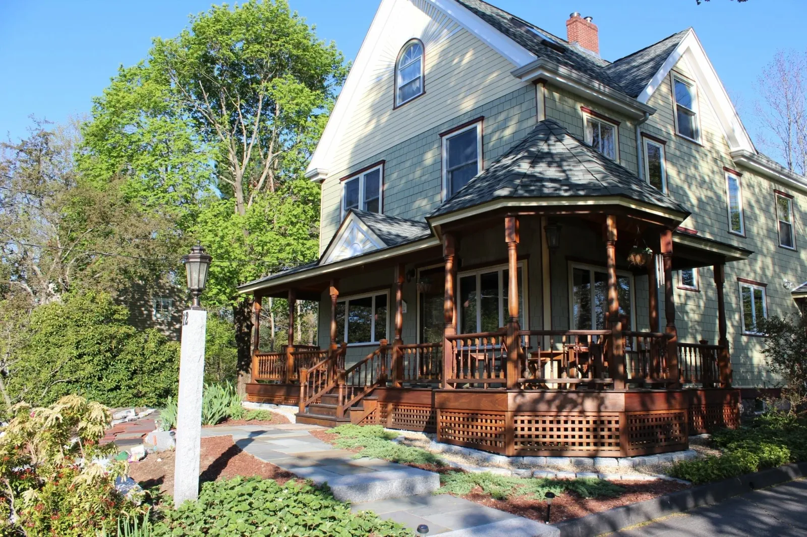 A three-story house with greenish siding, multiple windows, gable roofs, and a front porch with wooden railings and columns, surrounded by trees and a landscaped yard with a stone walkway.