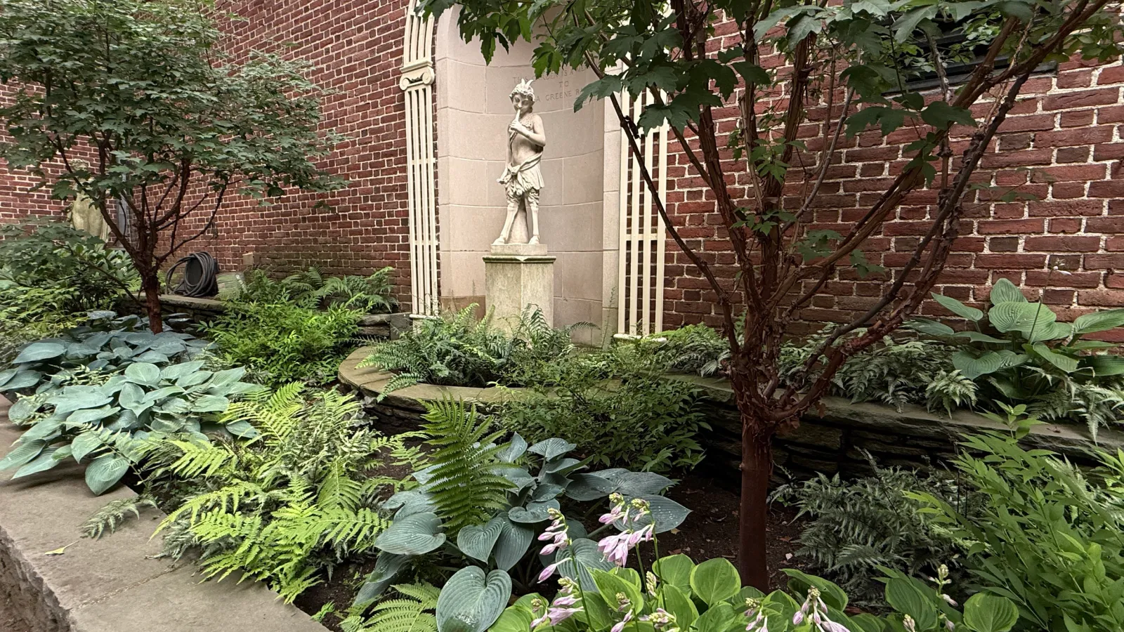 A small garden with lush green plants, including ferns and hostas, in front of a red brick wall. There is a white sculpture of a woman in front of a brick wall with a plaque. A garden hose is coiled on a black holder.