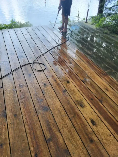 Person pressure-washing a wet wooden deck beside water, with a hose and coiled cord visible.