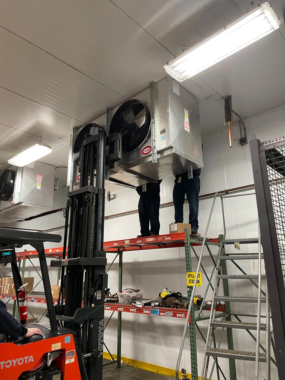 A warehouse scene with two workers standing on a shelf, repairing or installing large fans attached to the ceiling. A forklift labeled 'Toyota' is visible in the foreground. Industrial lighting and shelving with tools are in the background.