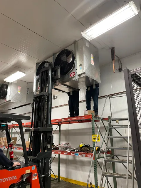 A warehouse scene with two workers standing on a shelf, repairing or installing large fans attached to the ceiling. A forklift labeled 'Toyota' is visible in the foreground. Industrial lighting and shelving with tools are in the background.