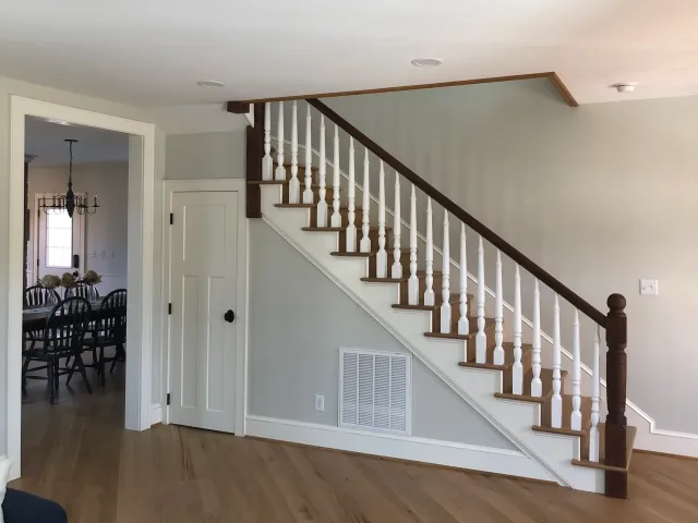 Interior view of a house showing a staircase with wooden handrail and white spindles, hardwood flooring, a door underneath the staircase, and a dining room with a chandelier and chairs visible through a doorway.