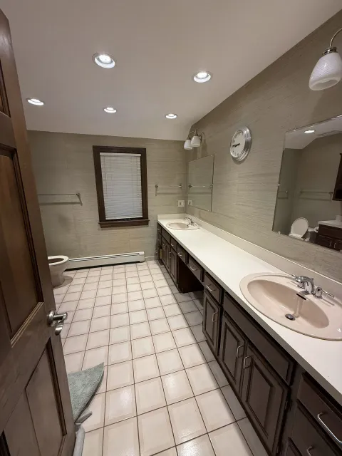 Bathroom with brown cabinets, two sinks, beige tile floor, textured wall, and a window.