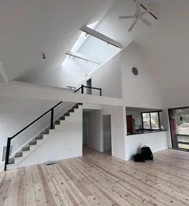 Interior of a modern, open-concept living space with a high ceiling, skylights, staircase, and large windows.