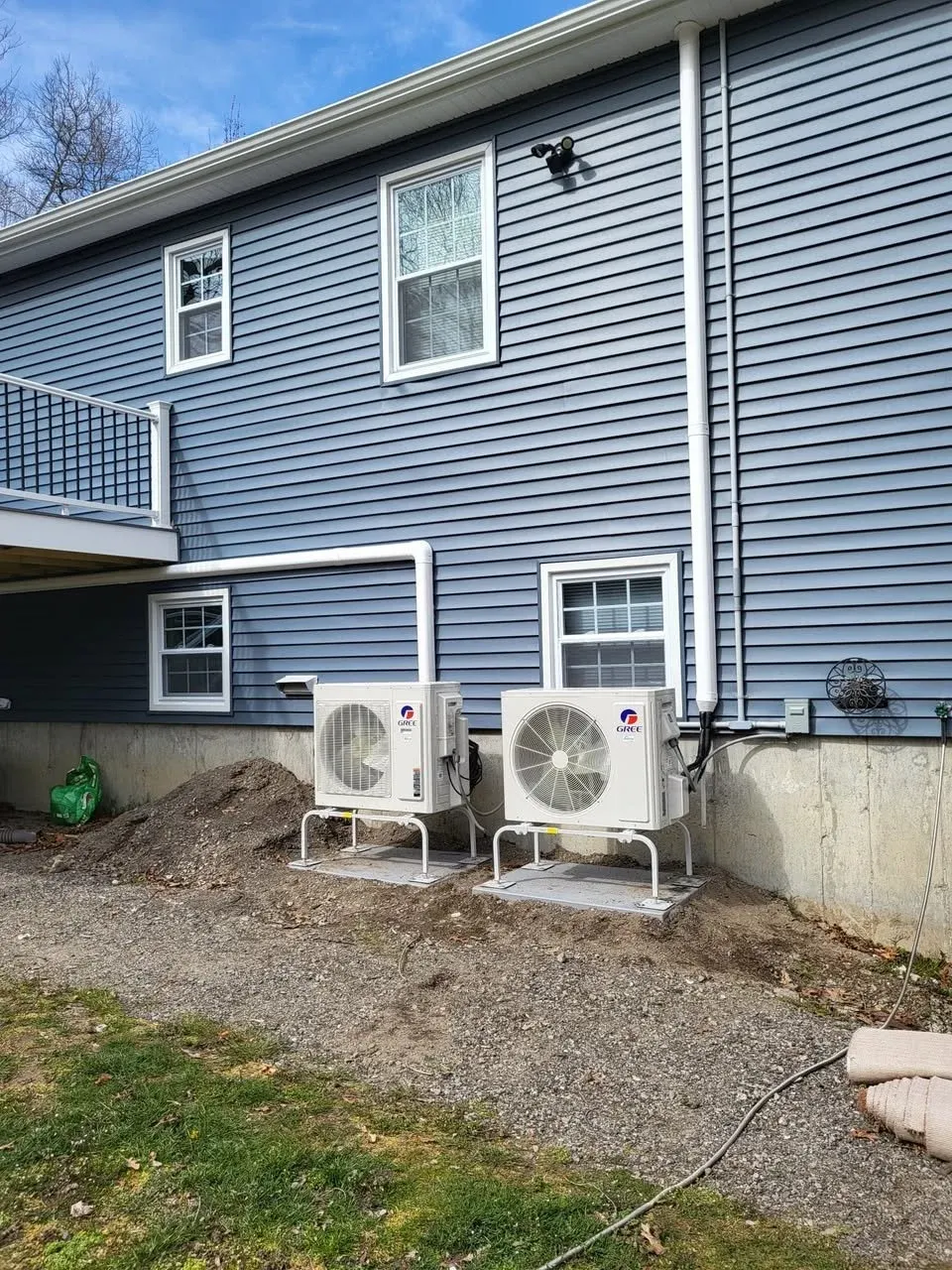 Two HVAC units on concrete pads outside a blue house with vinyl siding, windows, and a small deck.