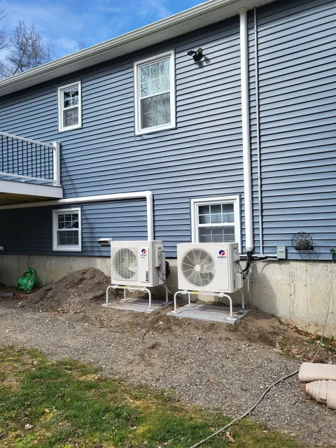 Two HVAC units on concrete pads outside a blue house with vinyl siding, windows, and a small deck.