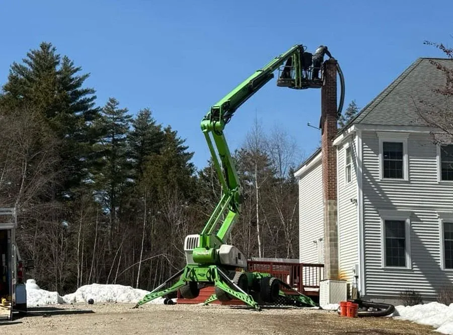 A green cherry picker extending to the chimney of a white house, with workers performing chimney maintenance or repair outside in a snowy yard.