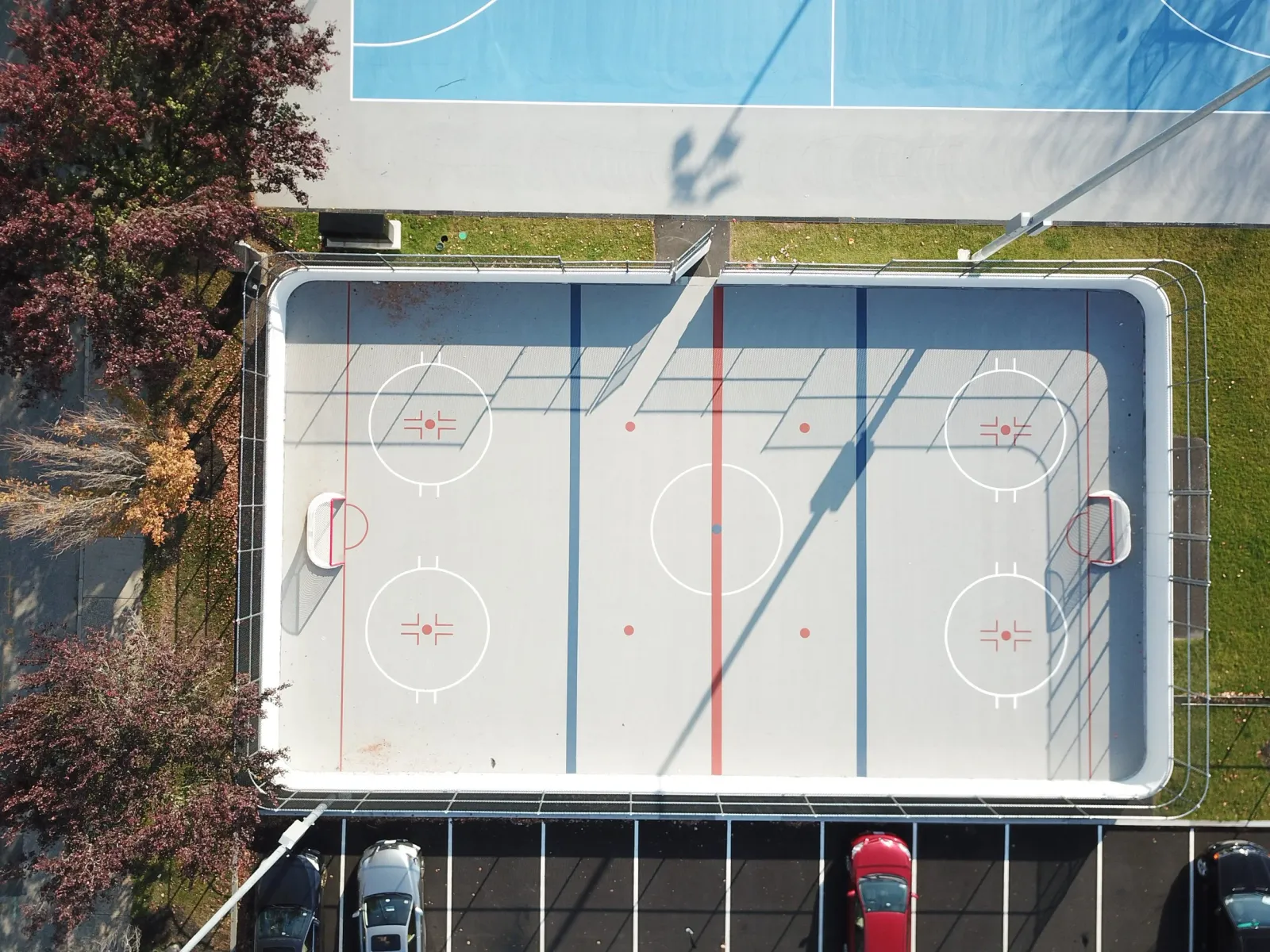 Aerial view of an outdoor hockey rink with red and blue lines, face-off circles, and goals at each end, surrounded by parked cars and trees with autumn foliage.