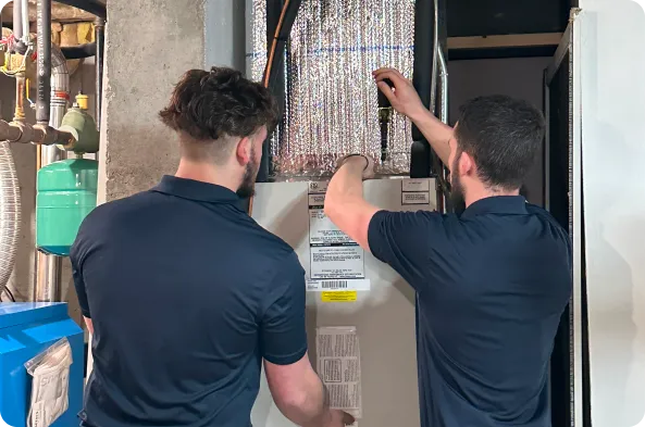 Two men in navy shirts perform maintenance on a hybrid heat pump in a utility room.