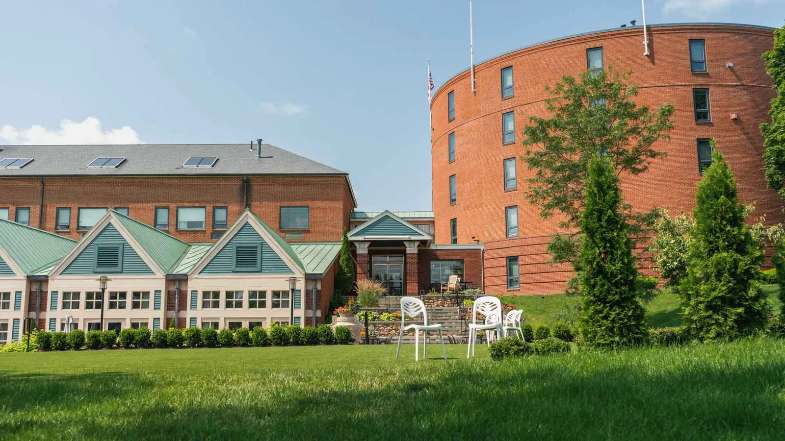 Brick building with green-roofed triangular sections and white chairs on a grassy lawn under blue sky.