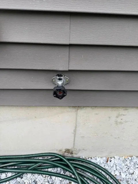 Outdoor wall faucet between gray siding and concrete foundation, with a green hose on the ground.