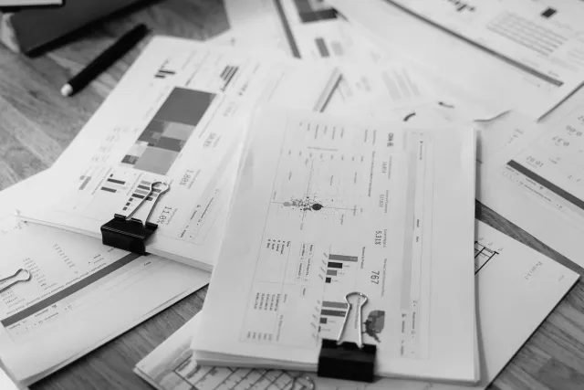Loose papers and documents spread out on a wooden table with some clips and a pen.